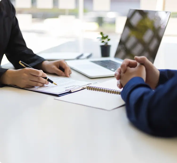 Two people in a professional meeting, one reviewing and signing documents across a desk from the other over credit report documents.
