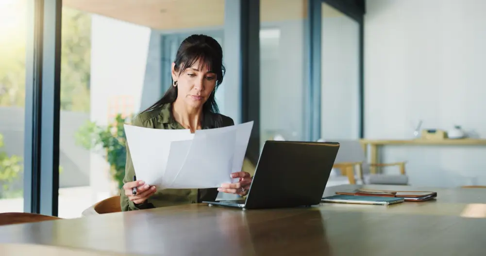 A woman reviewing documents at a desk with a laptop open in front of her.